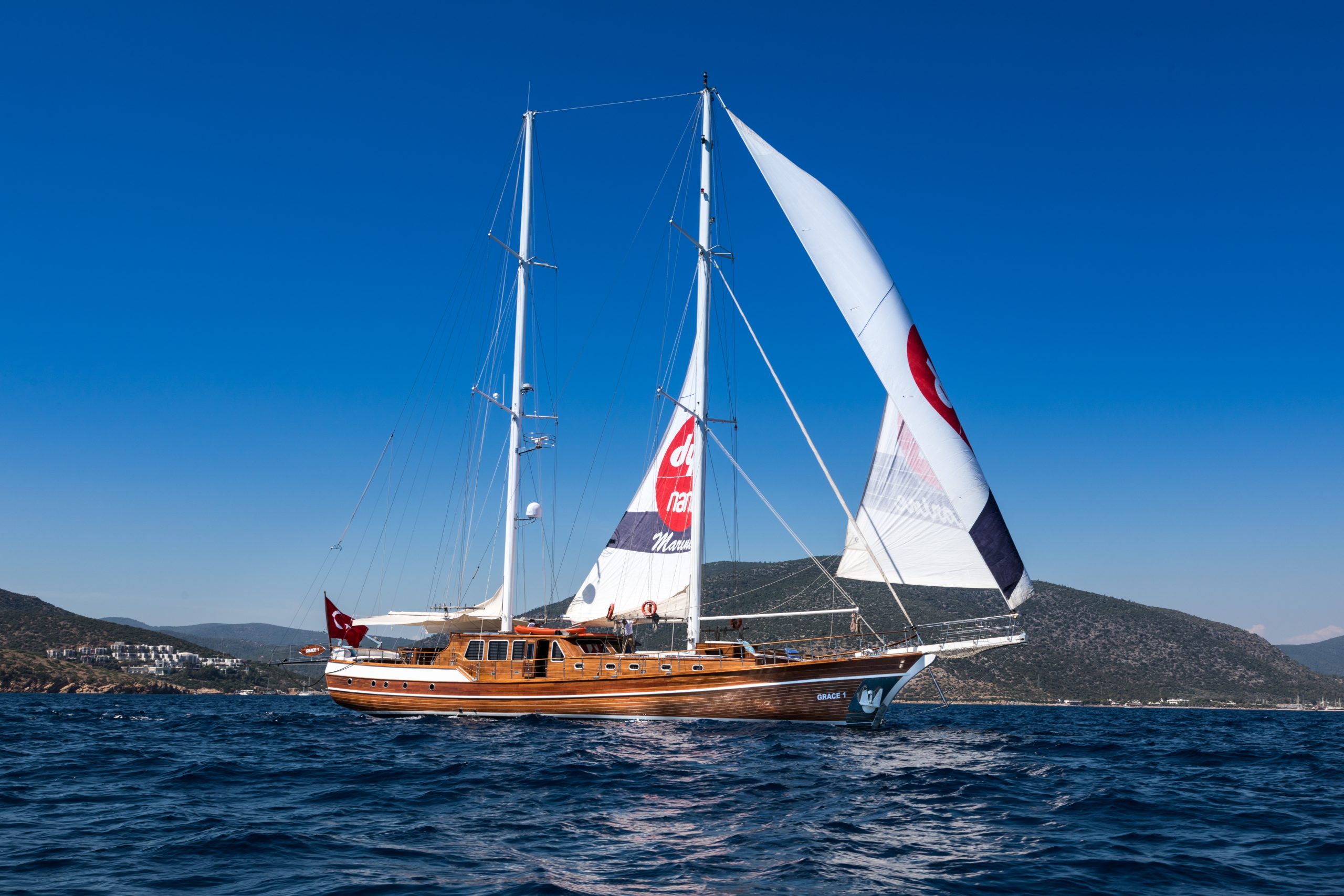 A large wooden sailboat with white sails glides on the blue sea under a clear sky, with green hills and distant buildings visible in the background.