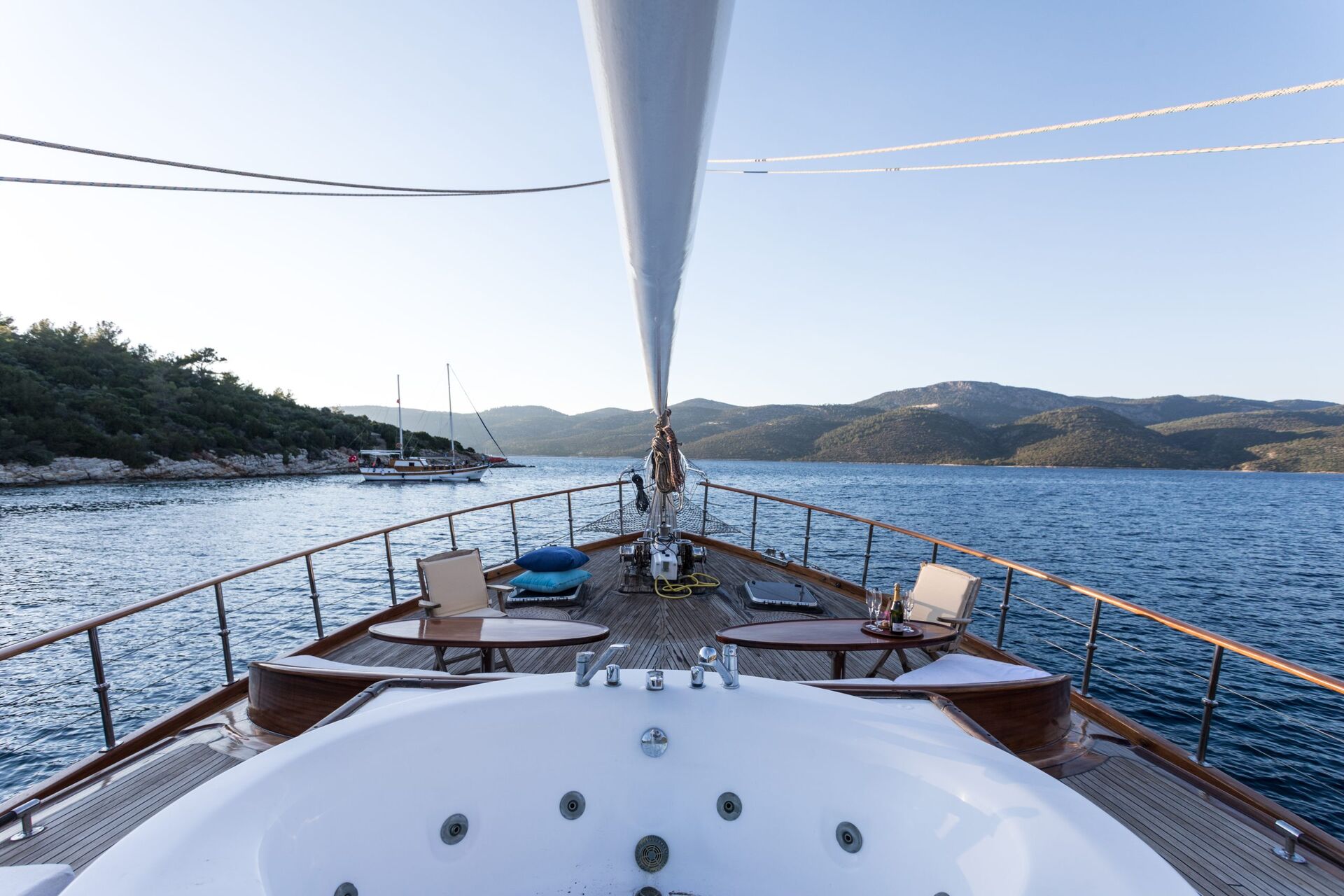View from the deck of a yacht featuring a jacuzzi in the foreground, lounge chairs, and a scenic backdrop of calm blue water with forested hills and anchored boats under a clear sky.