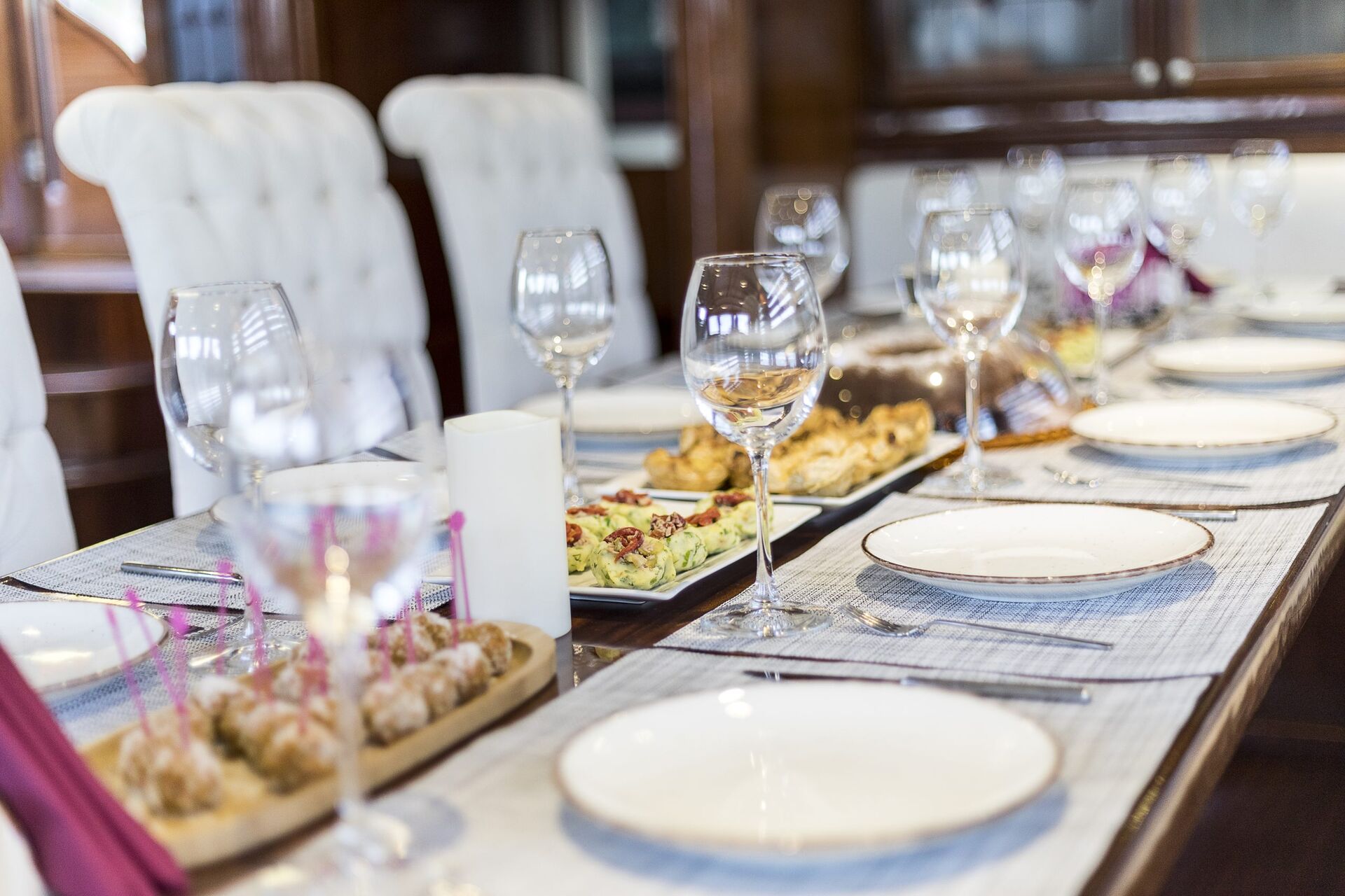 A dining table set with white plates, empty wine glasses, and various appetizers, ready for a meal in an elegant, well-lit room with white upholstered chairs.