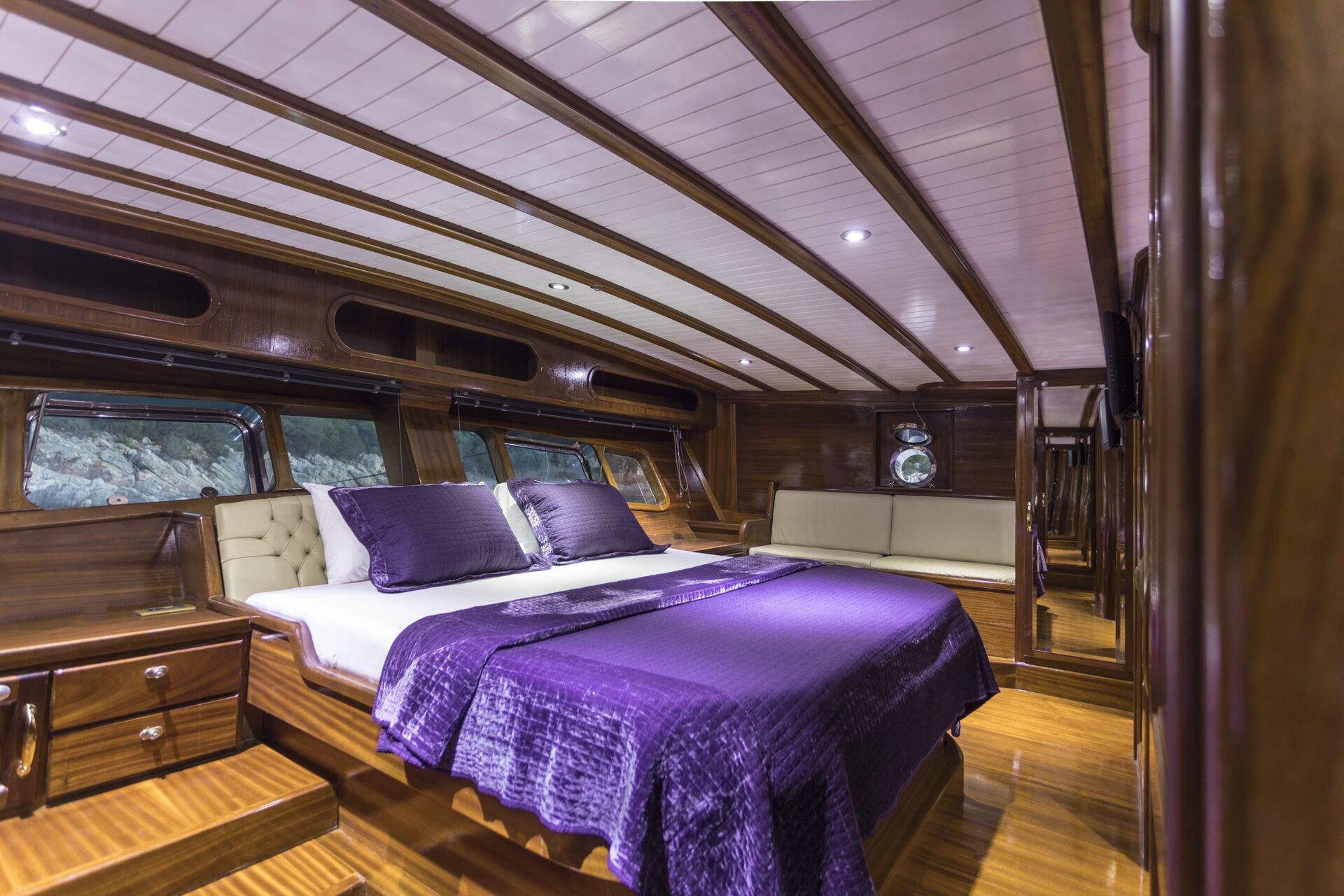 A luxurious yacht bedroom with polished wood walls and ceiling, a large bed with purple bedding and pillows, a beige cushioned bench, and windows offering a view outside. The room has a cozy, elegant ambiance.