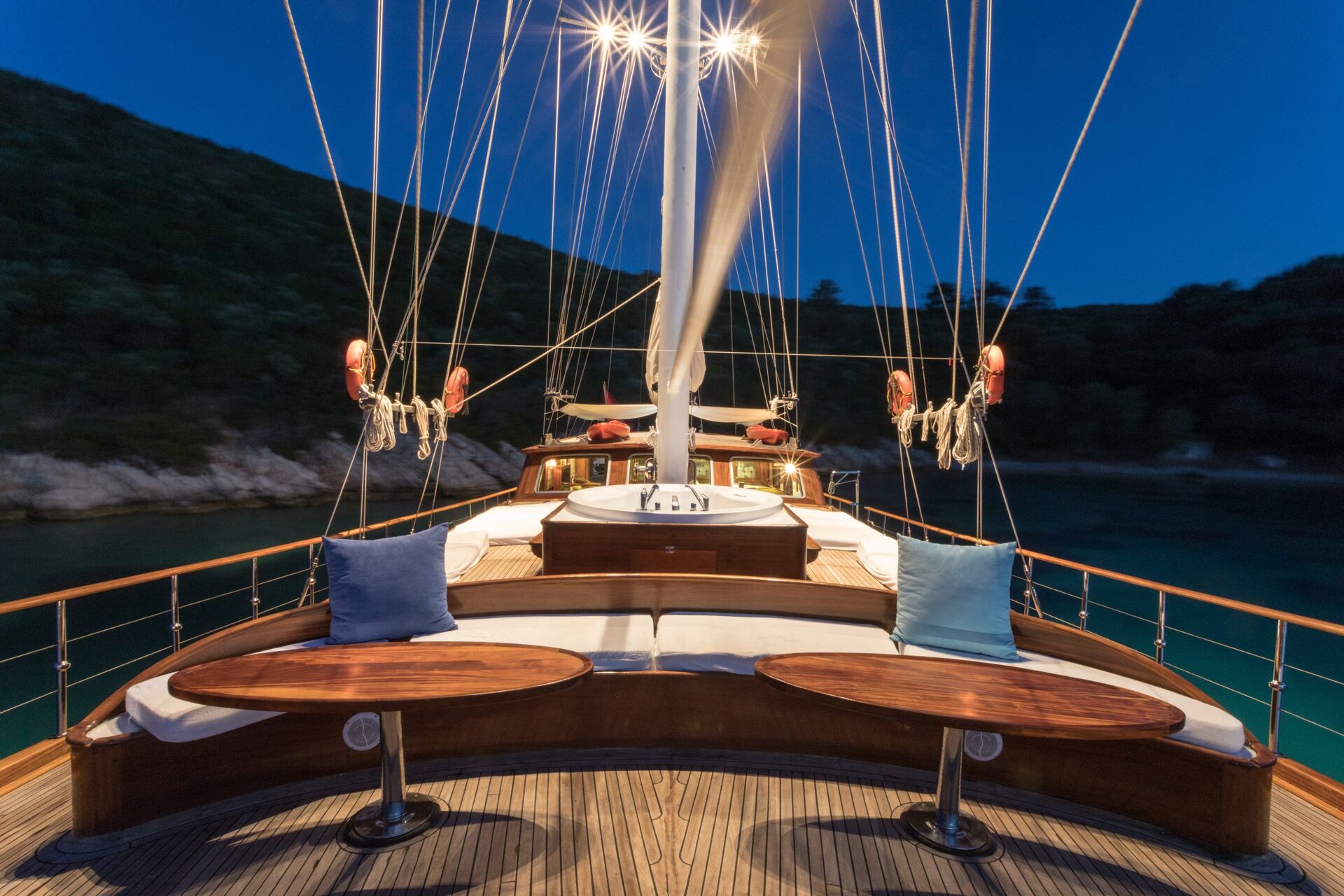 A wooden yacht deck at dusk, featuring curved benches with cushions and two polished tables. Ropes and masts rise above, with hills and calm water visible in the background under a deep blue sky.
