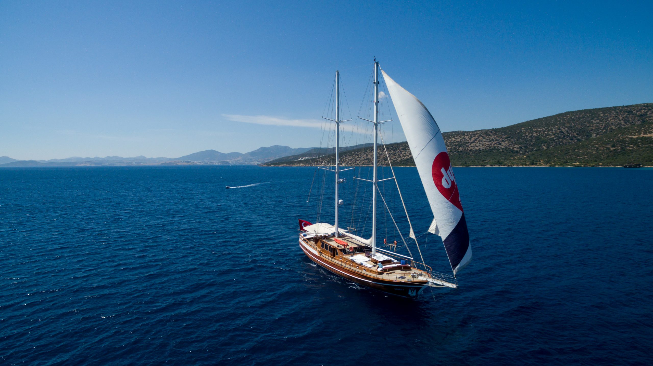 A large sailboat with a red-and-white spinnaker sails on deep blue water near a hilly coastline under a clear sky. Other boats and distant mountains are visible in the background.
