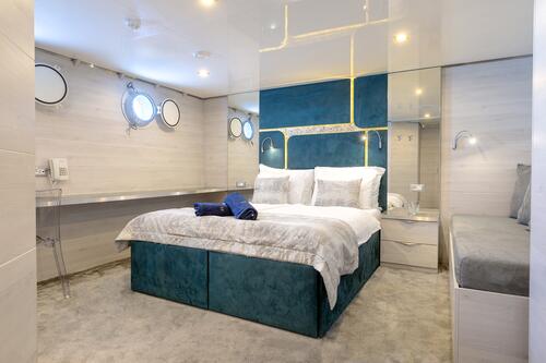 A modern bedroom with a teal upholstered bed, white and gray bedding, mirrored walls, porthole windows, a floating desk with a clear chair, folded towels, and a built-in bench on a light gray carpet.