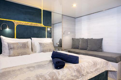 Modern hotel room with a neatly made double bed, two navy towels on the blanket, a built-in headboard with gold accent lighting, a gray cushioned bench, and white paneled walls.