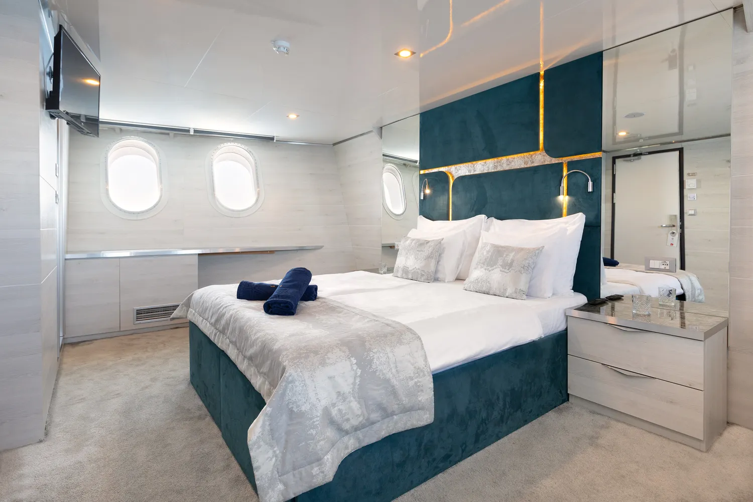 Modern bedroom with a large bed, white and silver bedding, and blue upholstered headboard. Two porthole windows, bedside tables, lamps, a wall-mounted TV, and neatly folded towels on the bed.