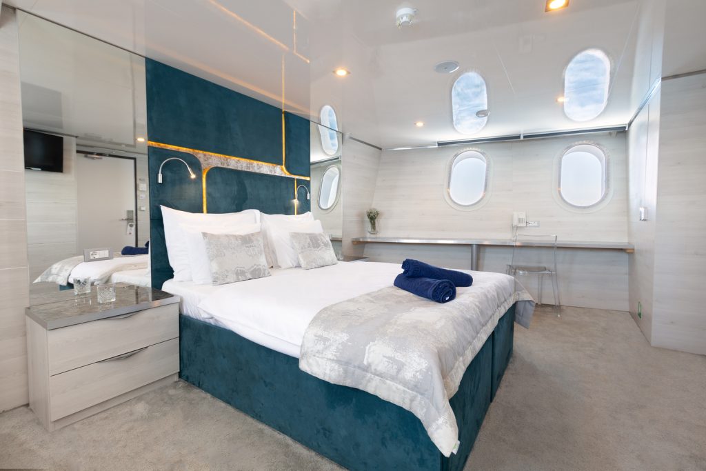 A modern, stylish bedroom with a large bed, blue and white bedding, bedside tables, porthole windows, a long desk, and soft lighting, creating a cozy and luxurious atmosphere.