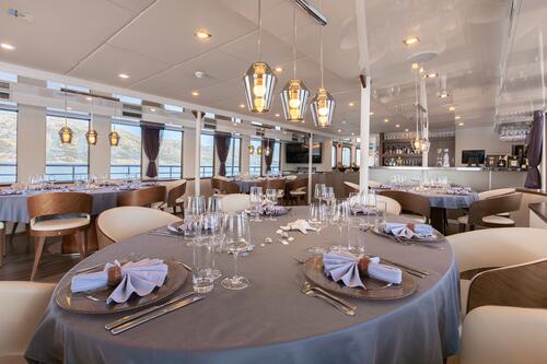 Elegant dining area with round tables set with glassware, plates, and folded napkins on gray tablecloths, modern wooden chairs, hanging pendant lights, and large windows with views of water and distant hills.