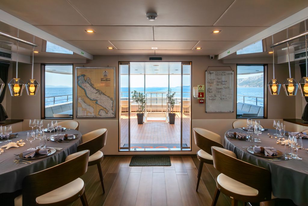 A modern yacht dining room with two round tables set for a meal, large windows showing the sea, a map on the left wall, a whiteboard on the right, and a view of a deck with plants and ocean beyond.