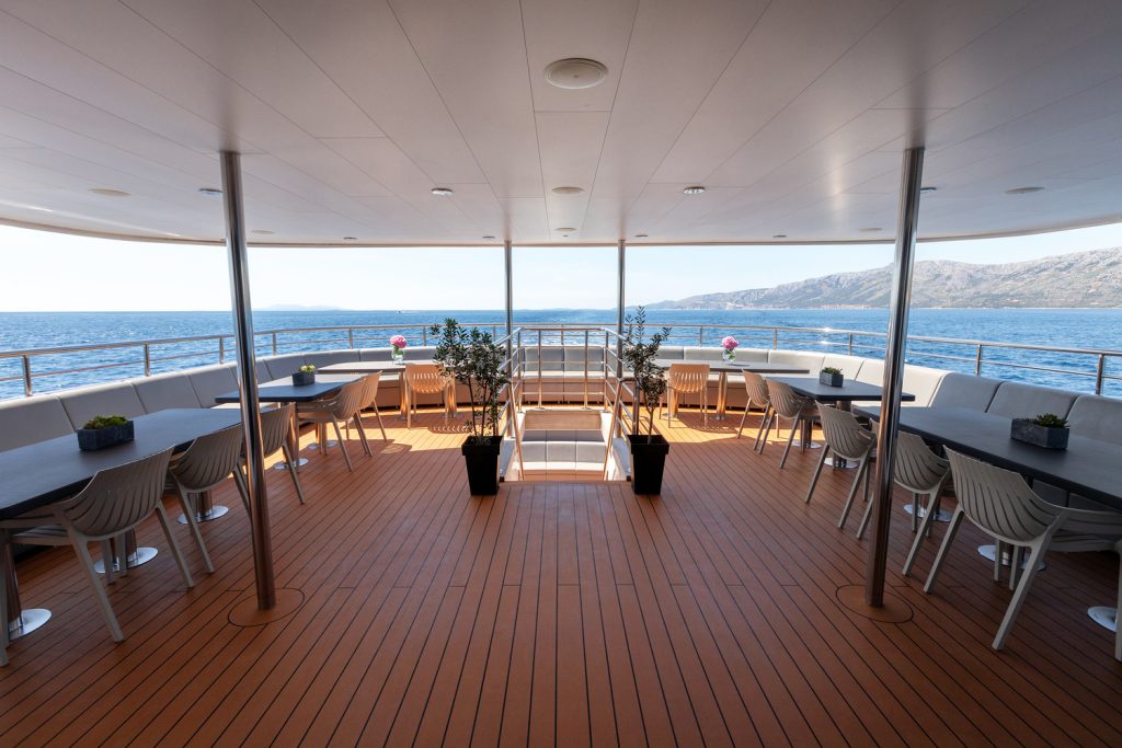 Spacious yacht deck with wooden flooring, modern tables and chairs, potted plants, and a clear view of the ocean and distant mountains under a bright sky.