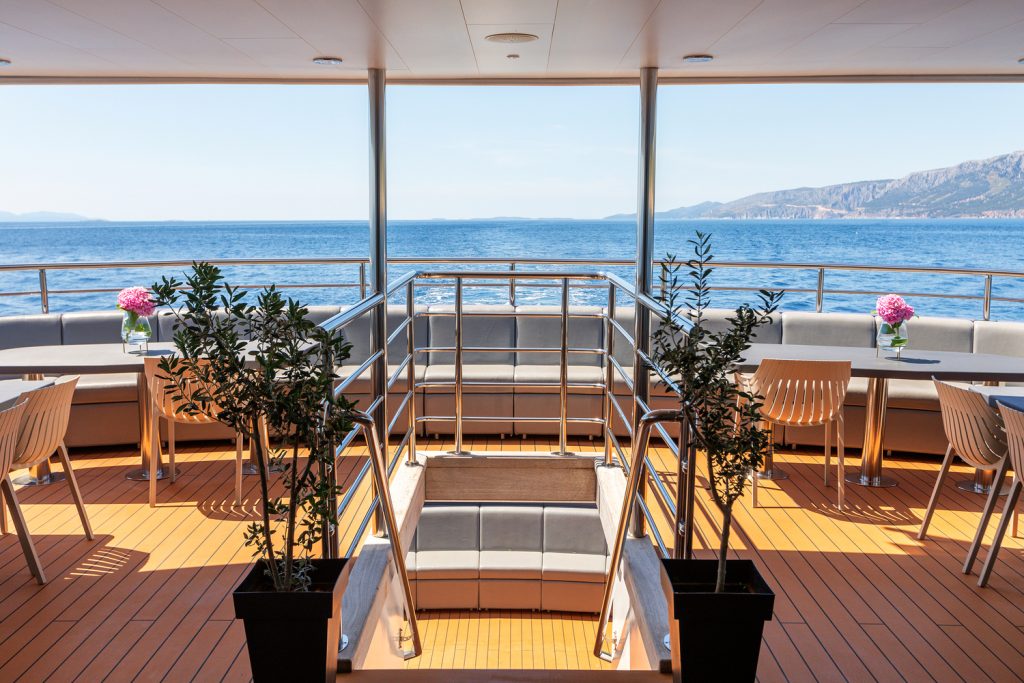Sunny yacht deck with tables, chairs, potted plants, and pink flowers in vases. Metal stairs lead below deck. The ocean and distant mountains are visible in the background under a clear blue sky.