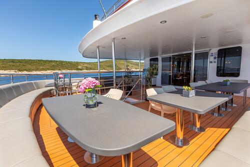 Outdoor dining area on a yacht with modern tables, chairs, potted plants, and cushioned seating, overlooking a calm blue sea and green coastline under a clear sky.