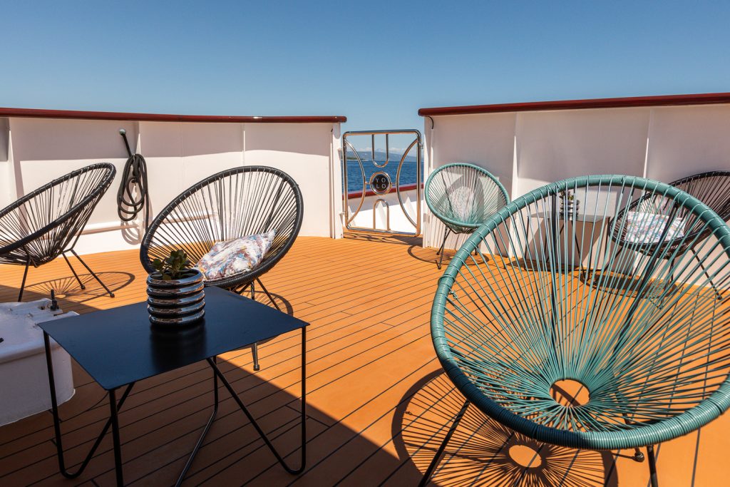 A sunny ship deck with modern circular chairs and a black table, overlooking the blue ocean under a clear sky. A nautical wheel is visible on the ship’s railing in the background.