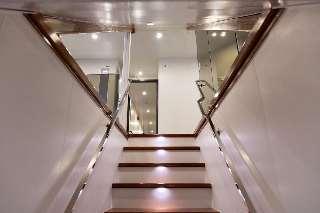 A modern staircase with wooden steps and built-in lights leads upward, flanked by glass railings and white walls, reflecting the bright lights in a sleek, contemporary interior space.
