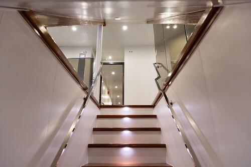 A modern staircase with wooden steps and built-in lights leads upward, flanked by glass railings and white walls, reflecting the bright lights in a sleek, contemporary interior space.