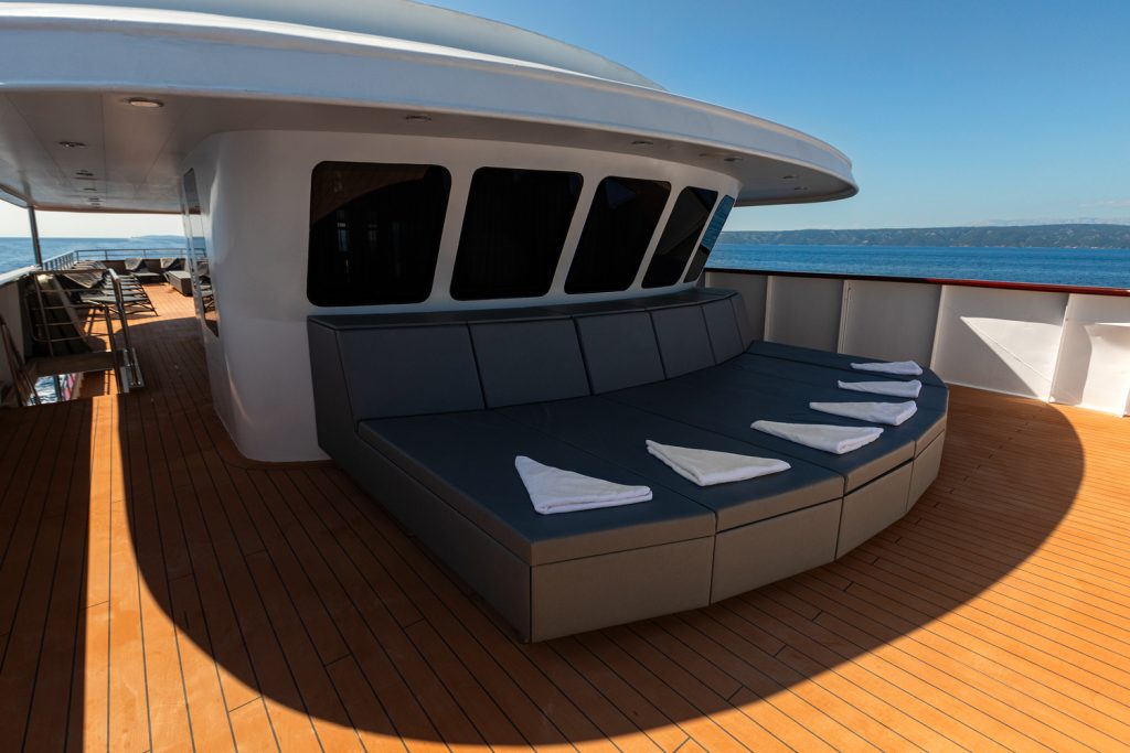 Modern yacht deck with a large cushioned lounge area, six folded towels placed on the seats, wood flooring, shaded canopy above, and ocean visible under a clear blue sky.