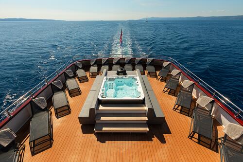 A luxury yacht deck with a central hot tub surrounded by lounge chairs, cruising on calm blue sea under a clear sky, leaving a white wake behind.