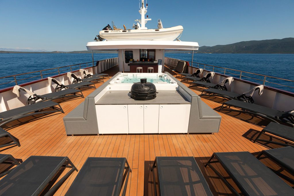 Sun deck of a yacht with multiple lounge chairs, a central hot tub, and a bar area at the front. The yacht is on calm blue water with distant green hills under a clear sky.