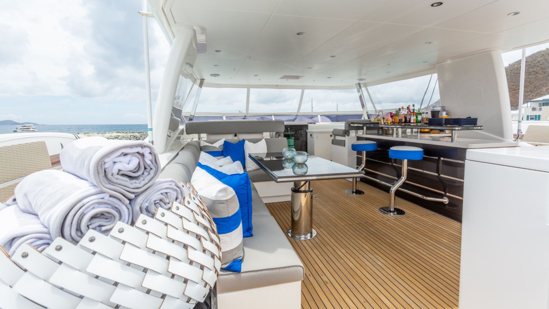 Modern yacht deck with cushioned seating, a table, rolled towels in a basket, blue bar stools, and a bar area. The deck overlooks the ocean and a cloudy sky, with some land visible in the background.