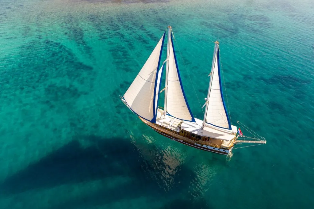 Aerial view of a wooden sailboat with two white sails floating on clear turquoise water, casting a large shadow on the sea below.