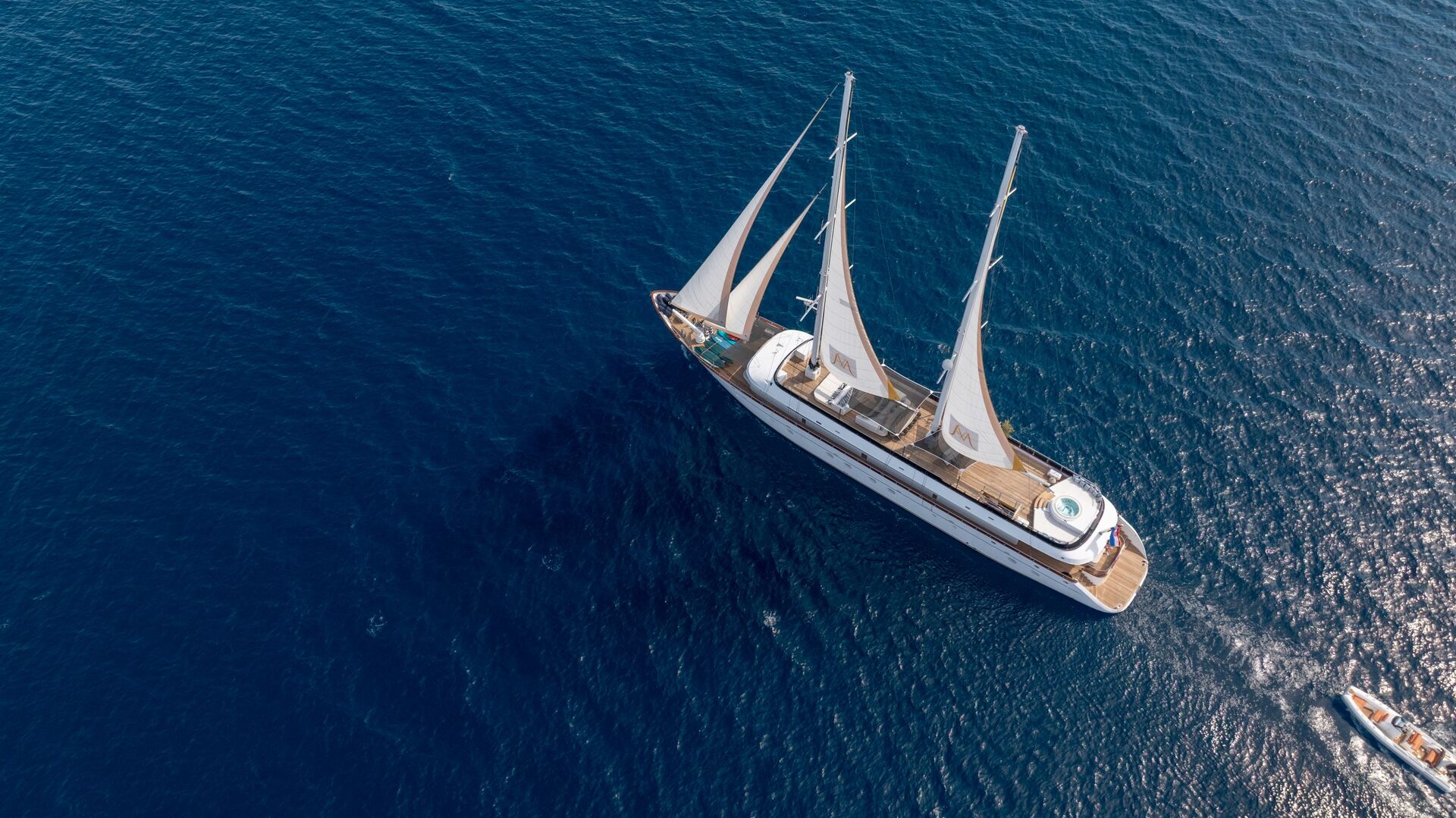 Aerial view of a large white sailboat with three sails gliding through deep blue water, accompanied by a small motorboat.