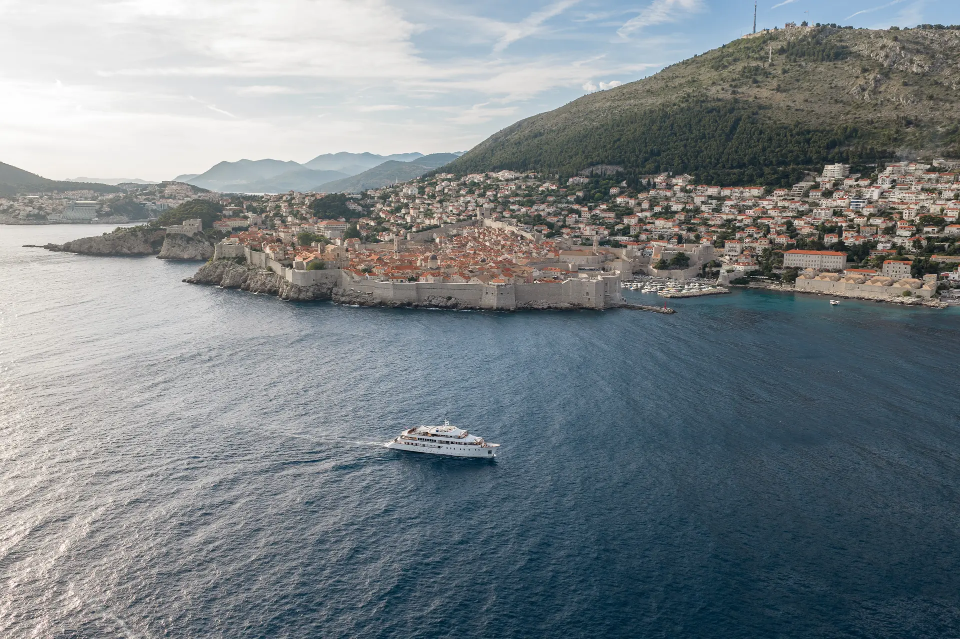 A large yacht sails near the historic walled city of Dubrovnik, Croatia, surrounded by blue sea with hills and houses in the background under a partly cloudy sky.