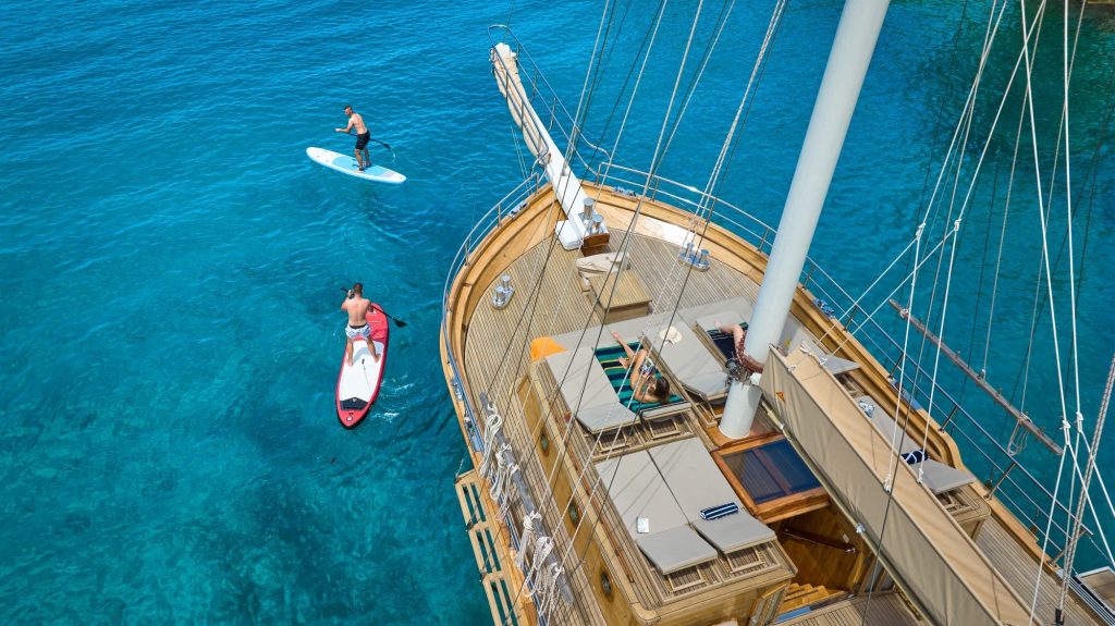 LIBRA Aerial view of two people paddleboarding near a large wooden yacht anchored in clear turquoise water, with sun loungers and seating areas visible on the yacht’s deck.