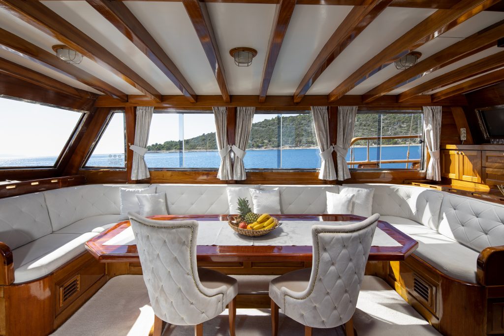 A luxurious yacht interior featuring a wooden table with fruit, surrounded by white cushioned chairs and a curved sofa. Large windows offer views of the sea and a distant hilly shore under clear skies.