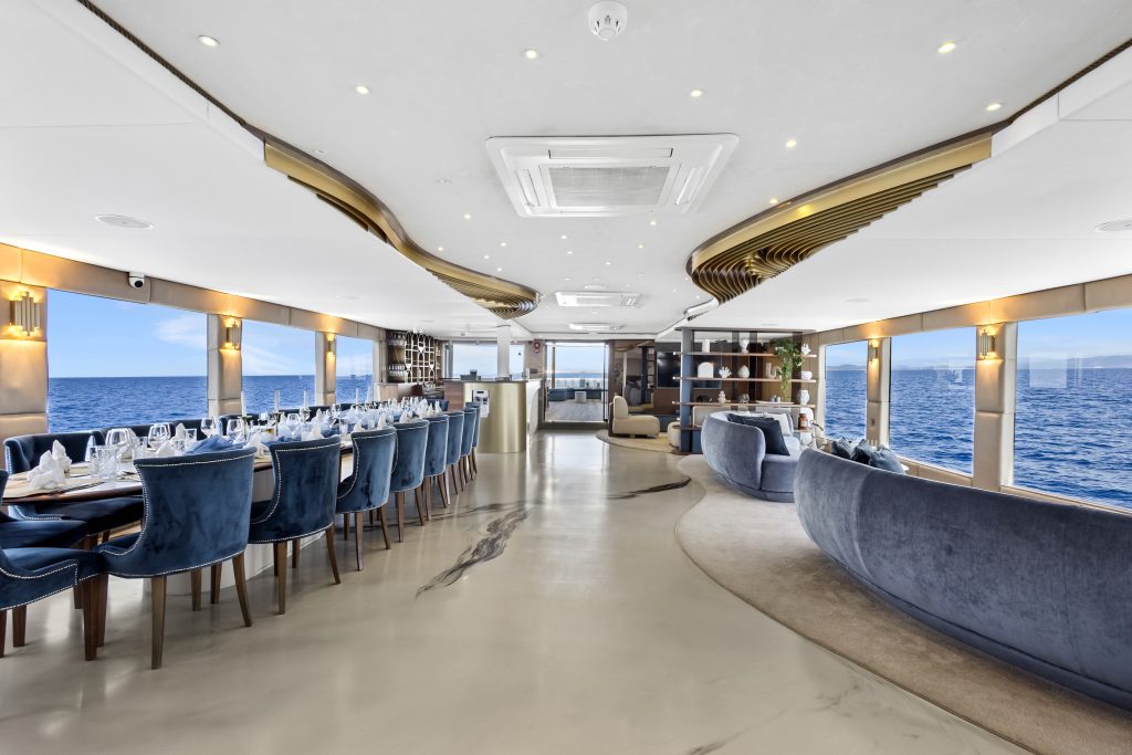 Elegant yacht interior featuring blue velvet chairs around a long dining table, curved sofas, large windows with ocean views, modern lighting, and stylish decor, creating a luxurious and spacious atmosphere.