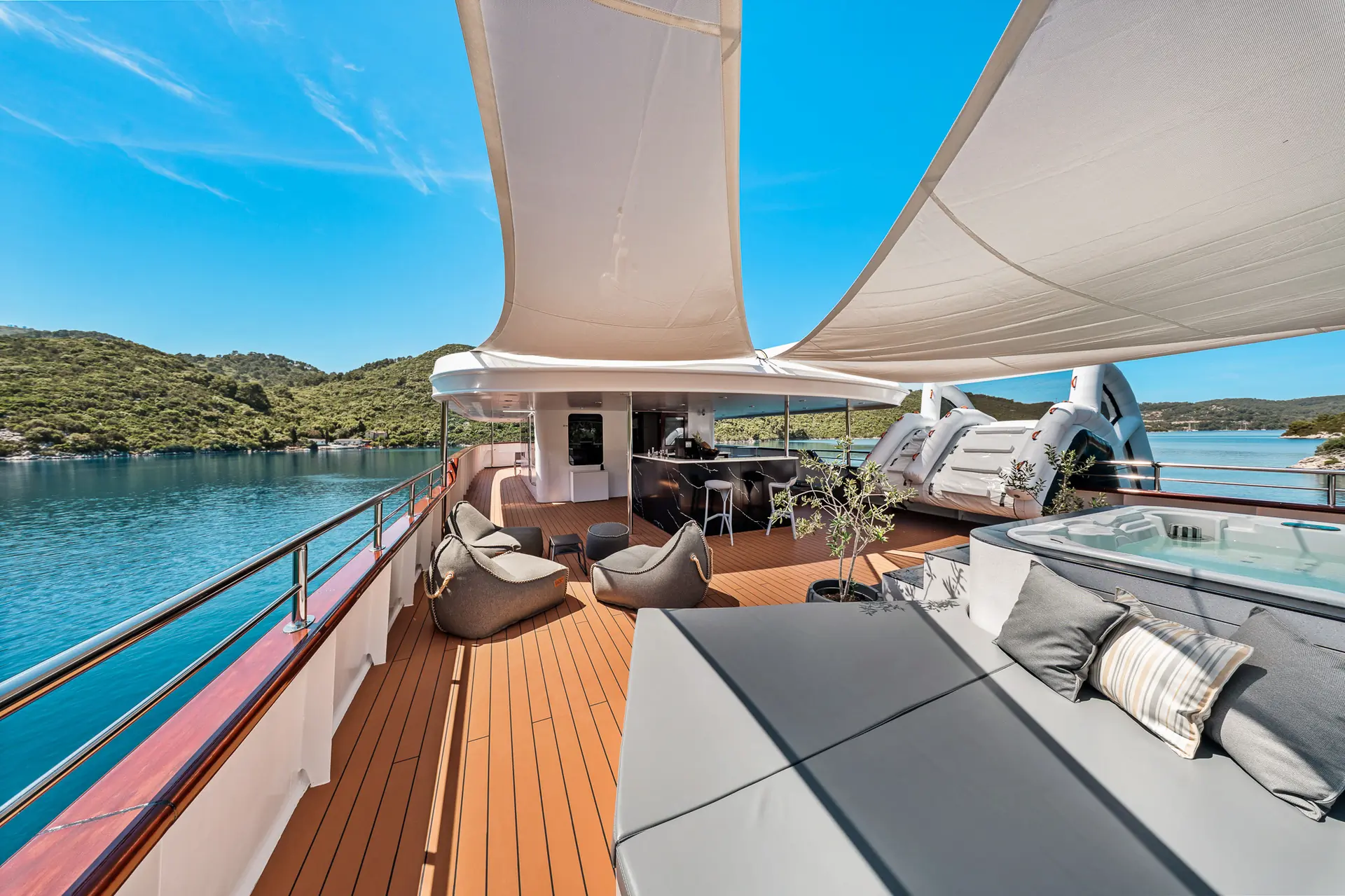 The deck of a luxury yacht features shaded lounge chairs, a hot tub, and outdoor seating, with views of calm blue water and green hills under a clear sky.