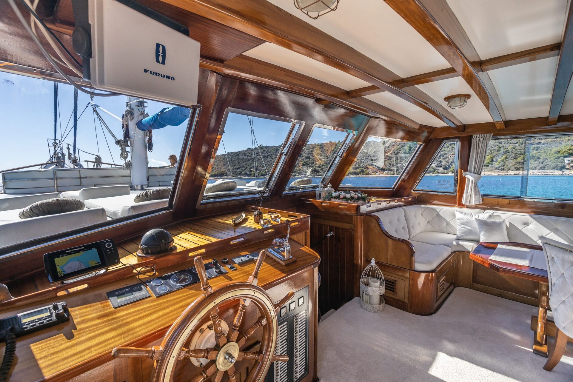 The interior of a wooden yacht cockpit with a large steering wheel, navigational equipment, and cushioned seating area. Sunlight streams in through wide windows overlooking blue water and a scenic shoreline.