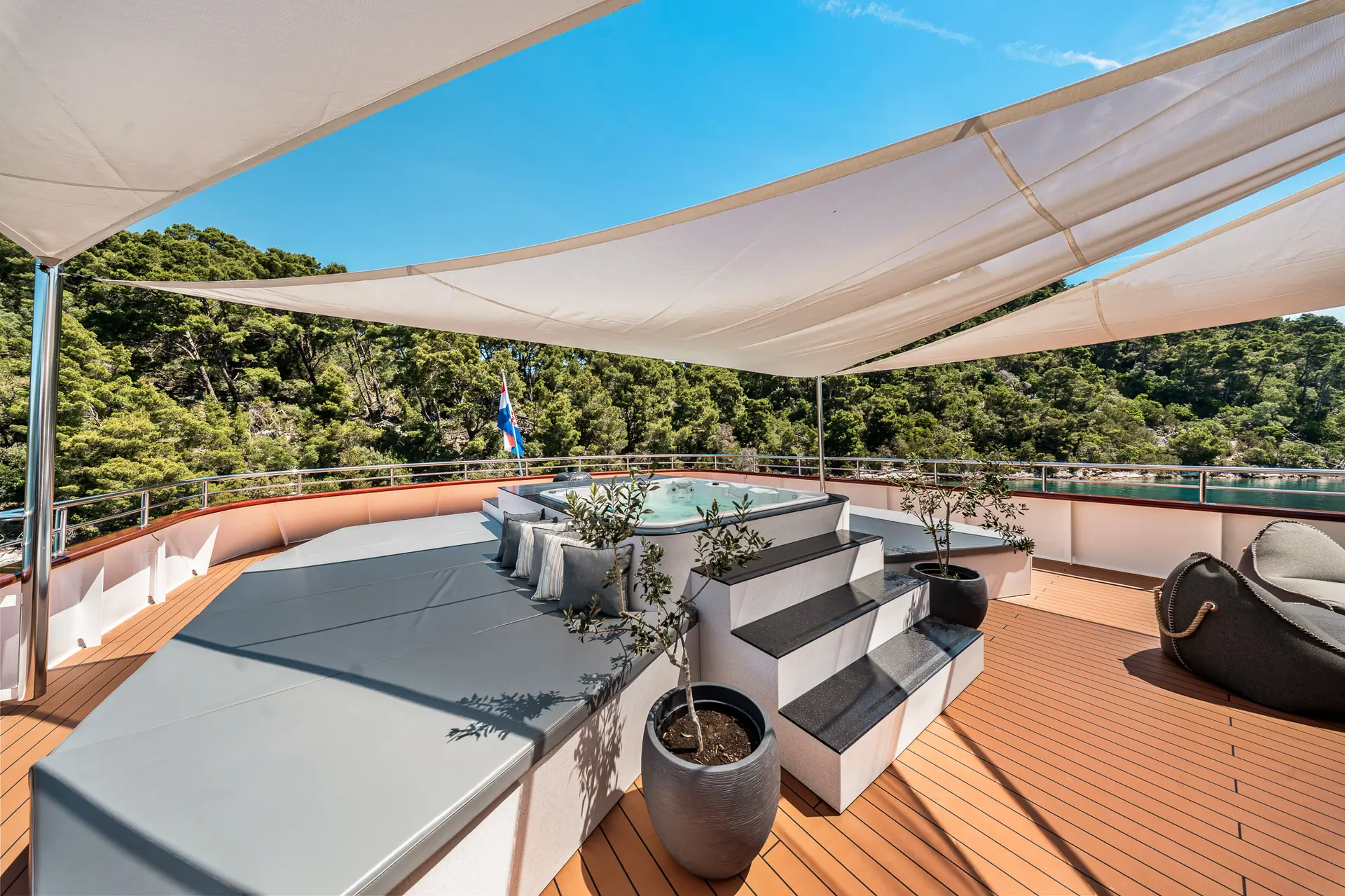 A luxurious yacht deck features a jacuzzi surrounded by padded seating, potted plants, and modern shade canopies. Lush green trees and blue sky are visible in the background.