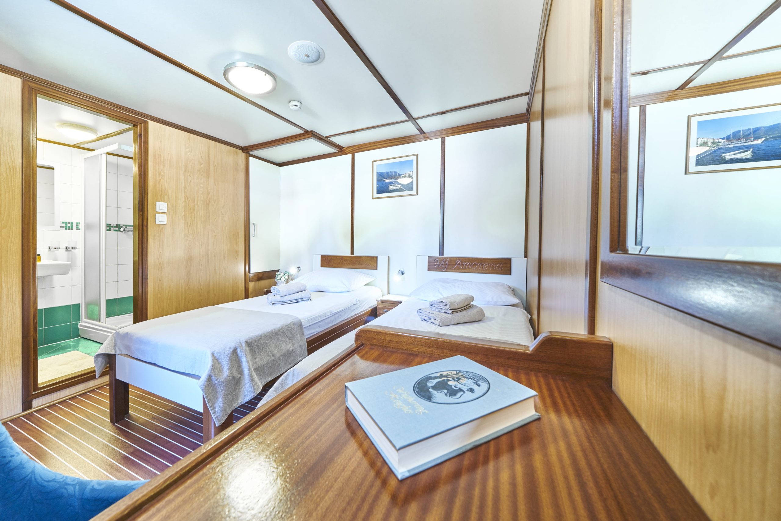 A bright cruise ship cabin with two single beds, wood paneling, a desk with a book, framed photos on the walls, and an open door revealing a bathroom with green tile accents—ideal comfort for your next yacht charter adventure.