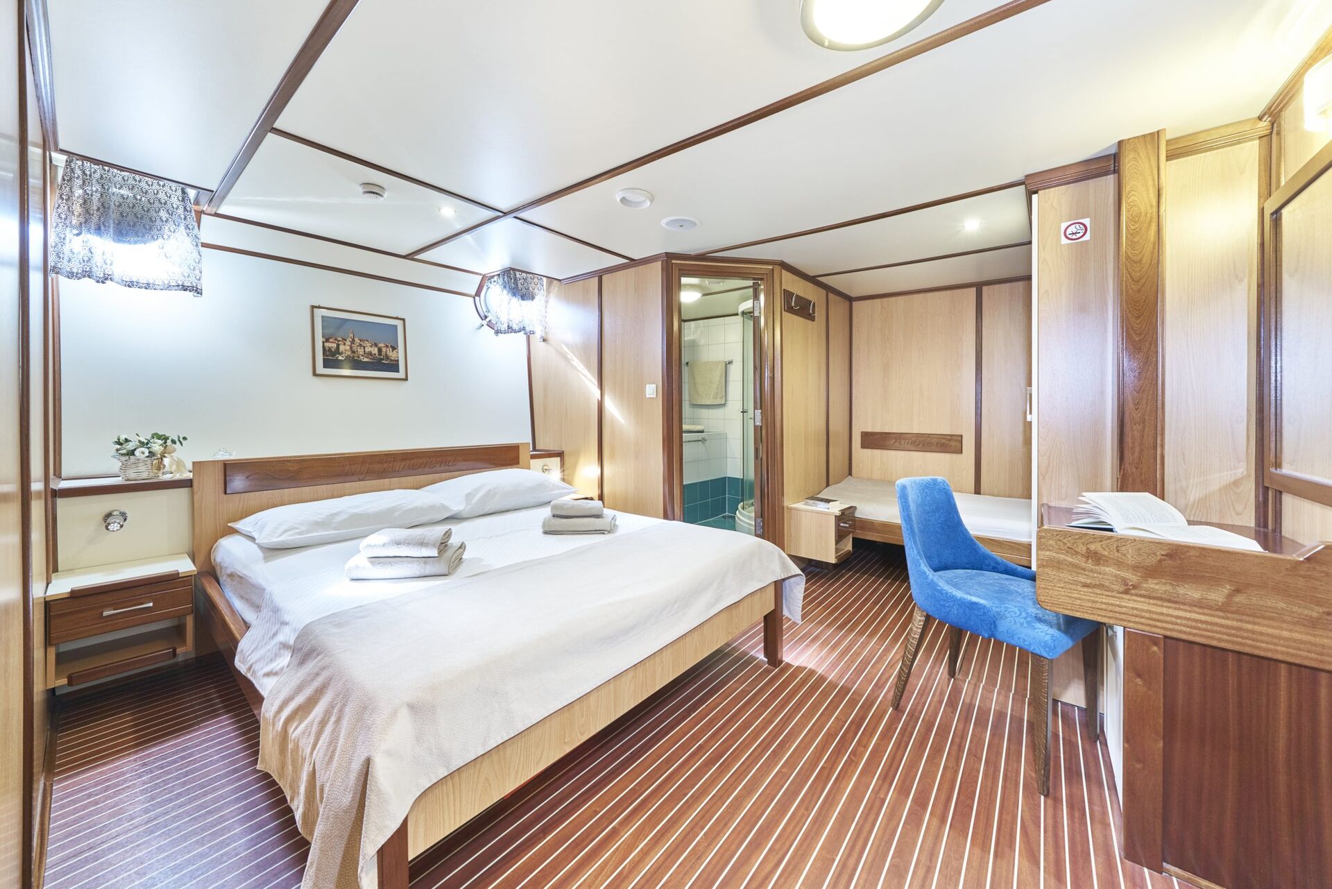 A bright, cozy cabin with a double bed, white linens, two towels, bedside tables, a small desk with a blue chair, wooden paneling, striped flooring, and an ensuite bathroom awaits you on your unforgettable yacht charter.