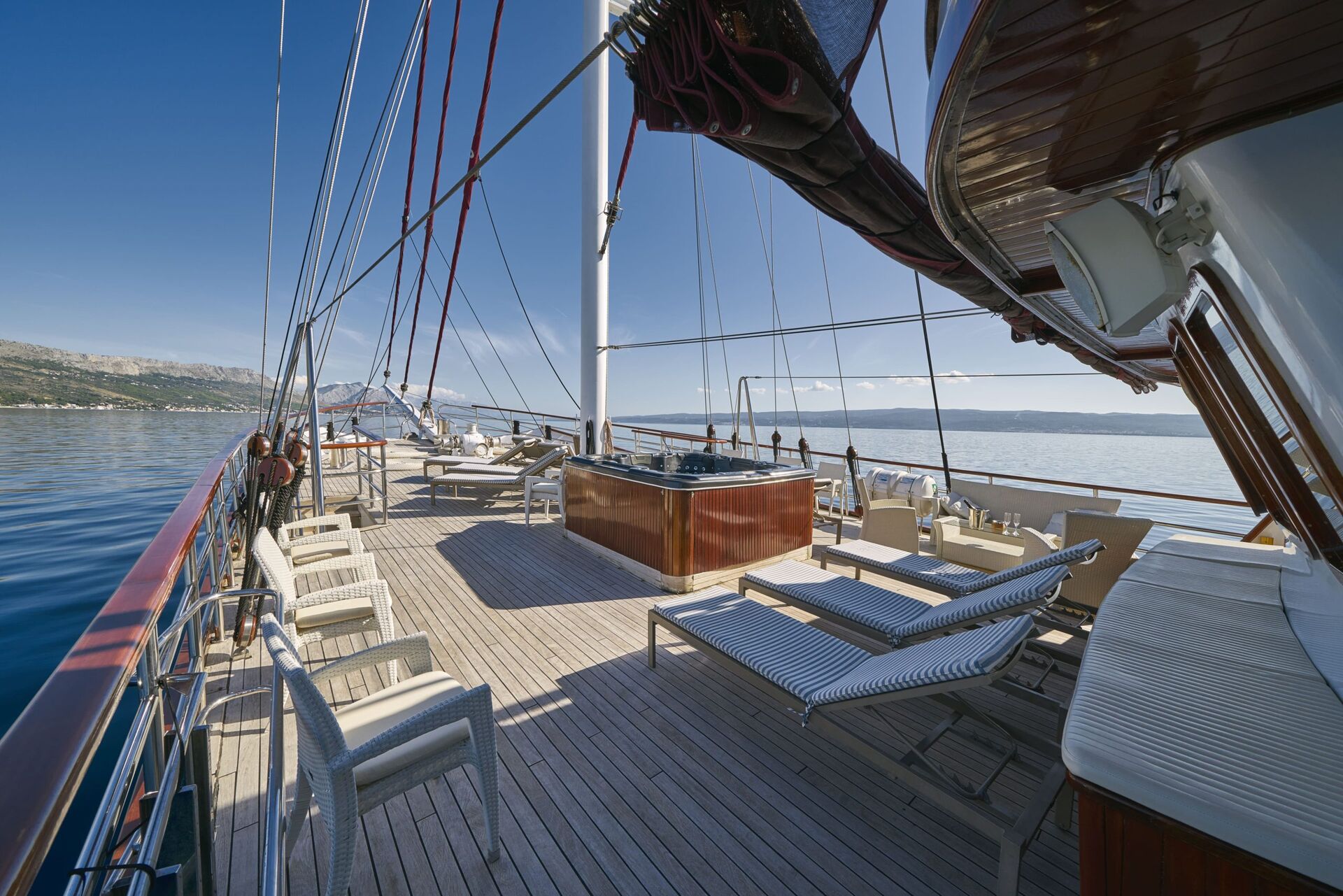 Sunny deck of a wooden yacht for charter with lounge chairs, hot tub, and seating area, overlooking the calm blue sea and distant shoreline under a clear sky.