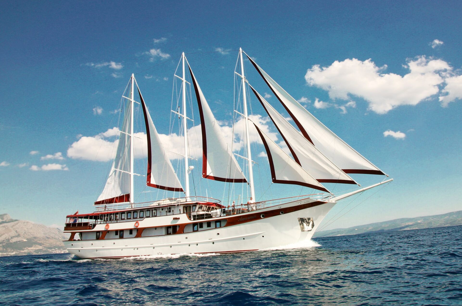 A large white sailboat with three tall masts sails on a blue sea under a bright, partly cloudy sky. This impressive yacht, ideal for yacht charter, has red accents, multiple decks, and distant land visible in the background.