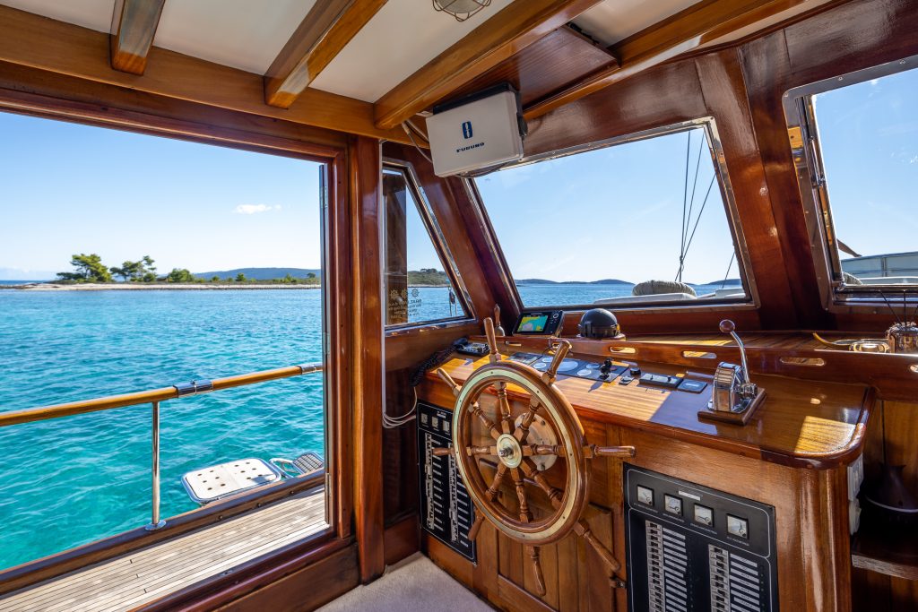 Wooden boat interior with a captain’s wheel, control panels, and windows offering a clear view of turquoise water, a nearby island, and blue sky outside. Sunlight illuminates the polished wood surfaces.