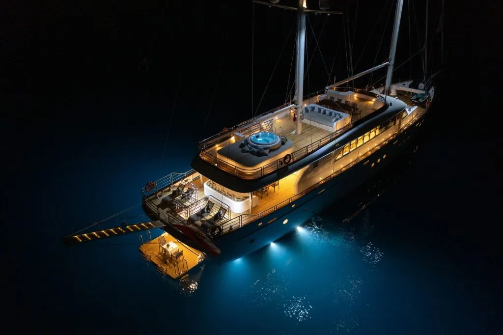 A luxury yacht is illuminated at night, floating on dark water. The yacht features a hot tub, outdoor seating areas, and underwater lights that cast a blue glow around the boat. A small dock is attached at the back.