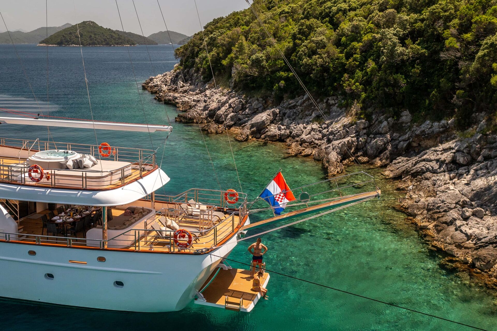 A luxury yacht with a Croatian flag is anchored near a rocky, tree-covered coastline in clear turquoise water. A person stands on the swim platform at the back of the yacht, enjoying the scenic surroundings.