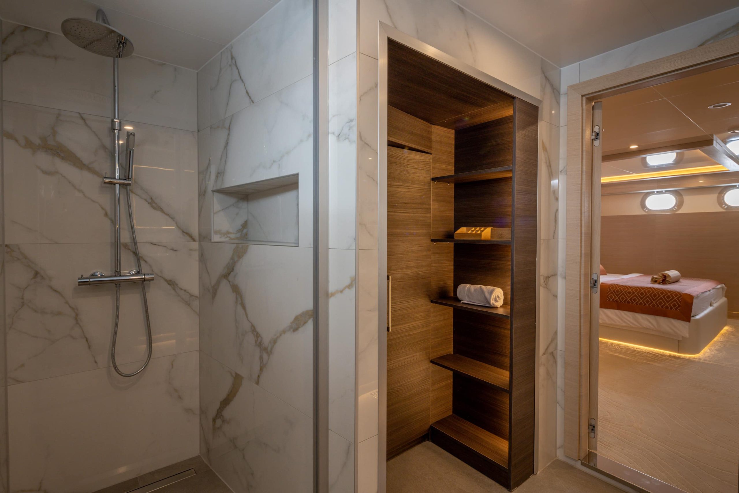 Modern bathroom with a marble-tiled shower on the left, a wooden shelving unit in the center holding towels and toiletries, and a bedroom with a bed and porthole windows visible through an open door on the right.