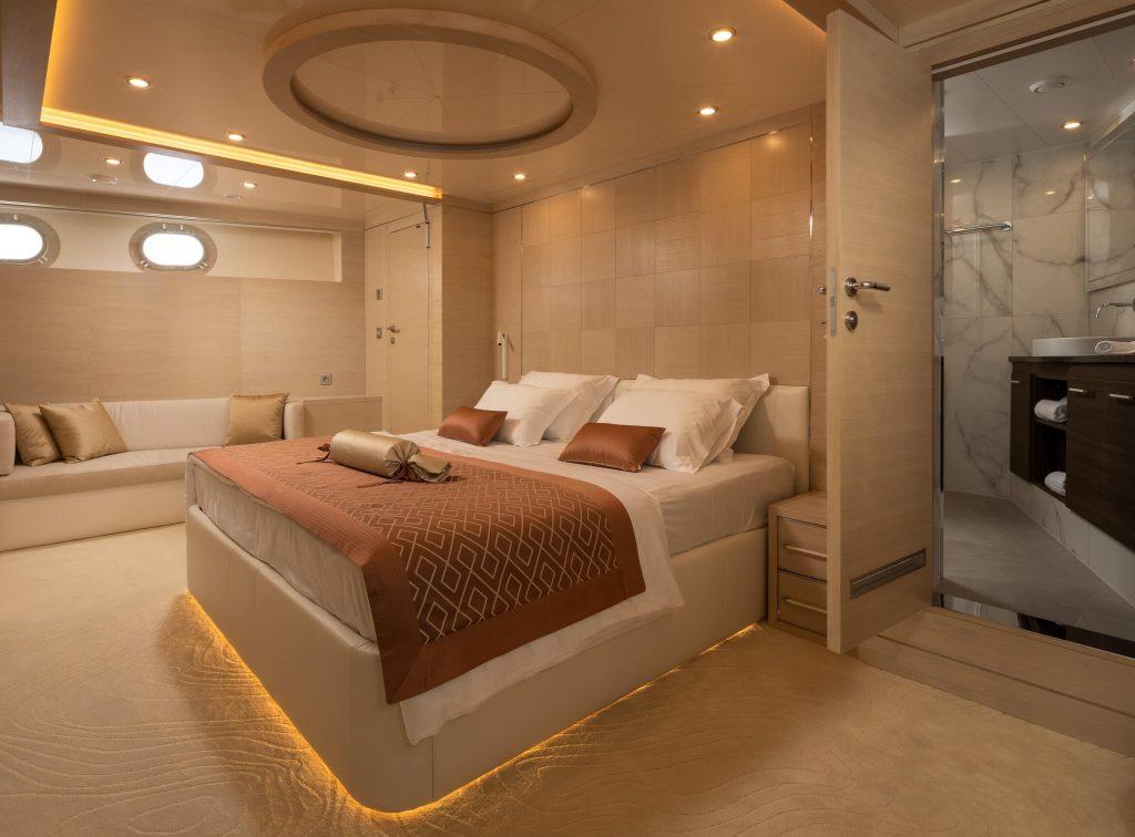 Luxurious yacht bedroom with a large bed, brown and white bedding, soft lighting, a circular ceiling feature, a cream sofa, porthole windows, and an open door leading to a modern marble bathroom.