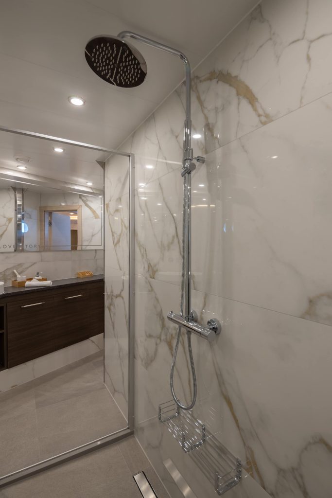 Modern bathroom with large marble tiles, a glass shower enclosure, chrome rainfall showerhead, and a wooden vanity with a mirror in the background. The space is well-lit with recessed ceiling lights.