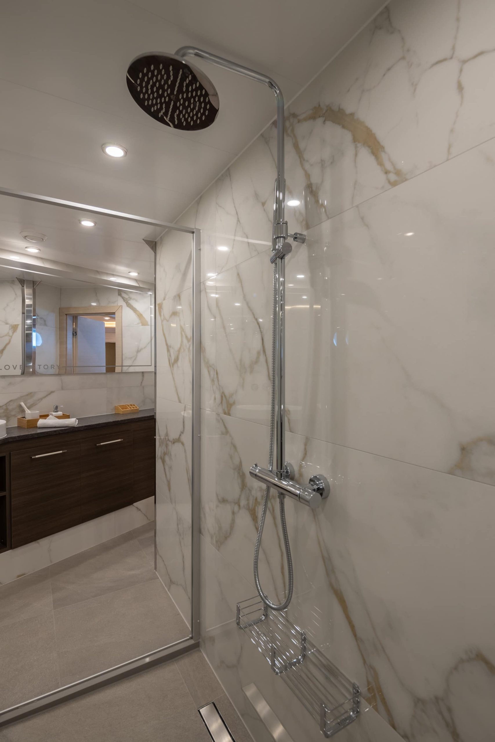 Modern bathroom with large marble tiles, a glass shower enclosure, chrome rainfall showerhead, and a wooden vanity with a mirror in the background. The space is well-lit with recessed ceiling lights.
