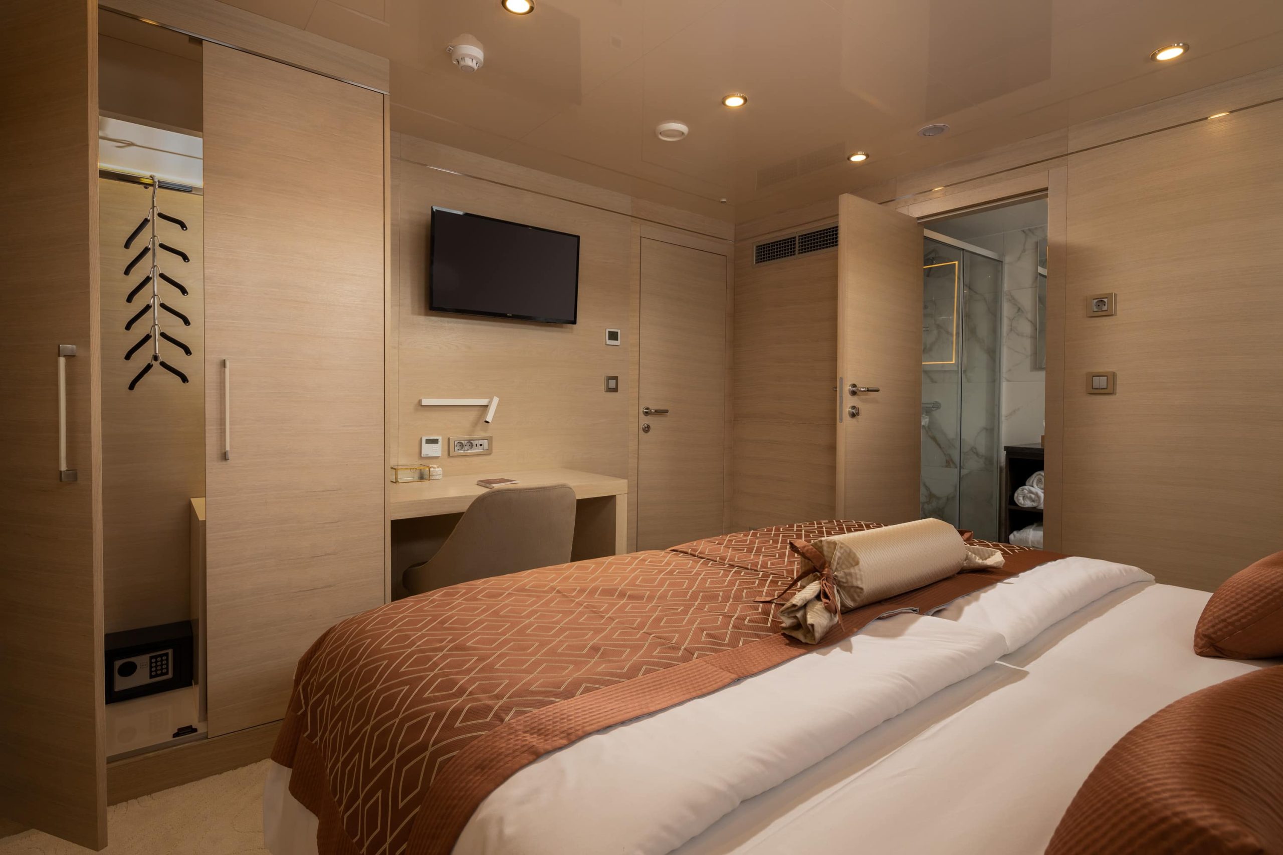 A modern hotel room with a double bed, brown and beige bedding, a desk with a chair, a wall-mounted TV, built-in wardrobes, and a door leading to a bathroom with marble walls.