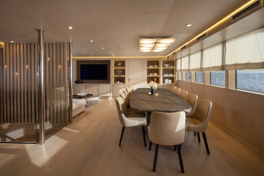 Modern yacht dining area with a long table and cream chairs, large windows with views of the sea, wooden floors, built-in shelves, and ambient ceiling lighting.