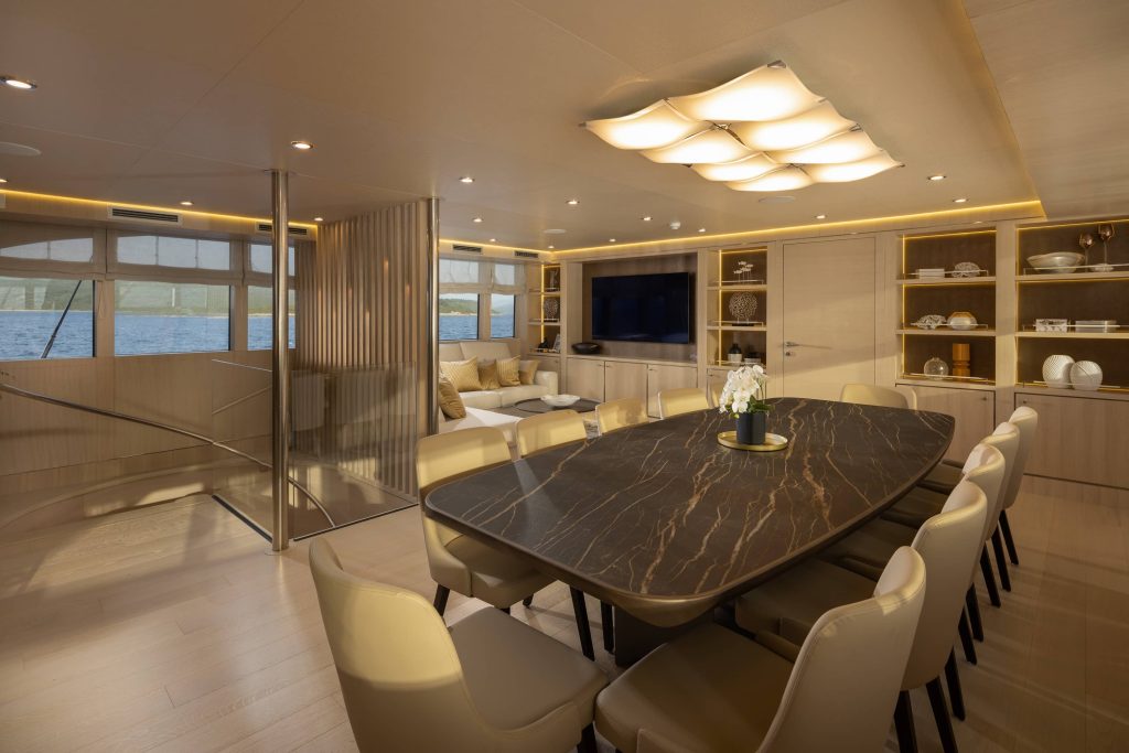 Luxurious yacht interior with a dark marble dining table surrounded by cream chairs, a modern spiral staircase, a cozy sitting area, shelves with decor, large windows showing water, and warm ambient lighting.