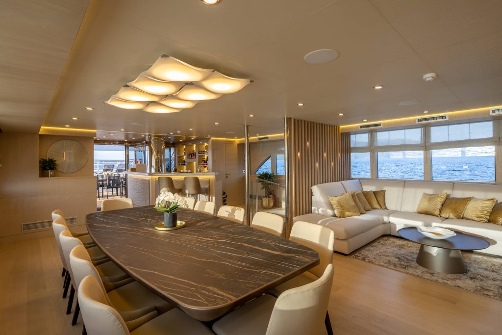 Luxurious yacht interior with a marble dining table, cream chairs, modern lighting, and a cozy white sectional sofa by large windows overlooking the sea. A bar and dining area are visible in the background.