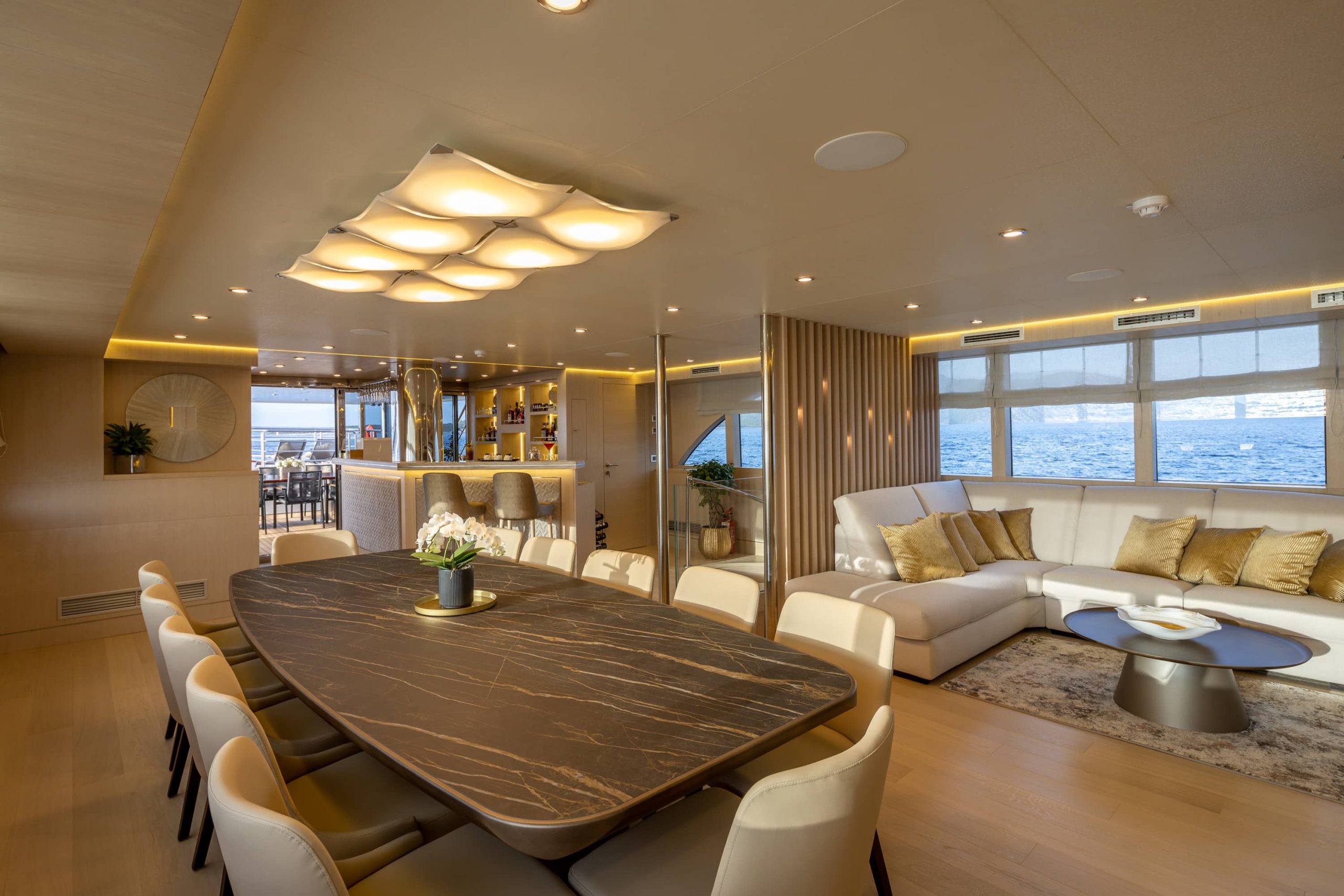Luxurious yacht interior with a marble dining table, cream chairs, modern lighting, and a cozy white sectional sofa by large windows overlooking the sea. A bar and dining area are visible in the background.