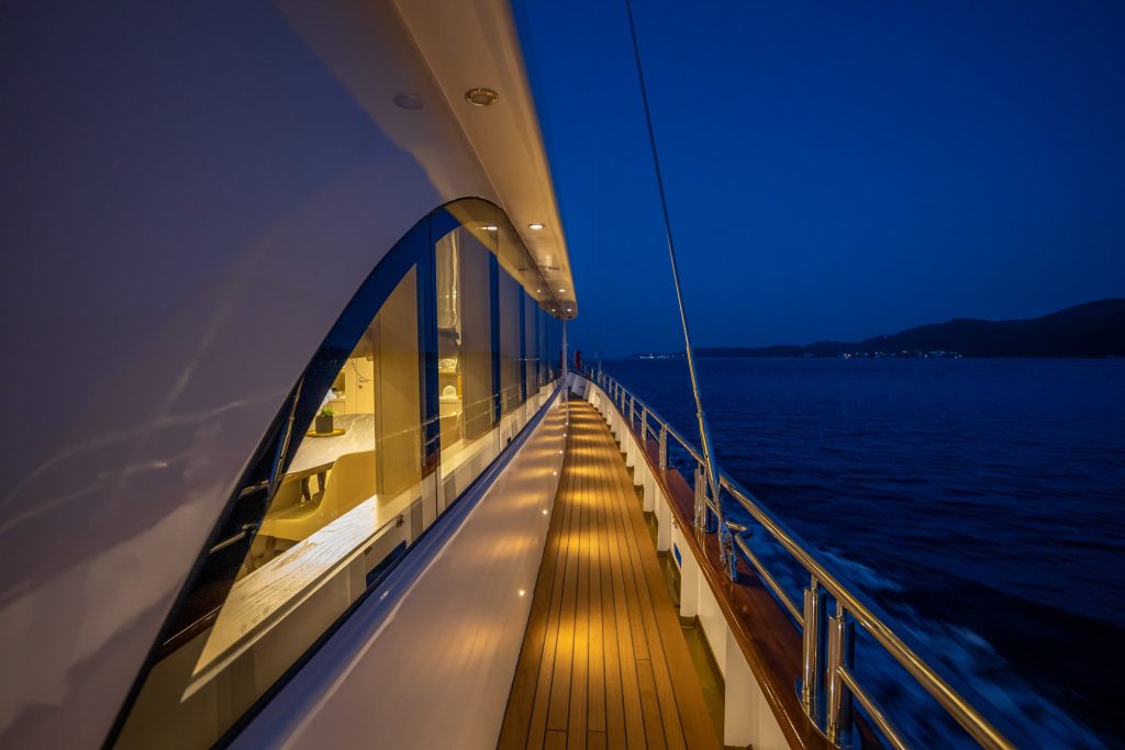 A yacht deck at night with warm lights illuminating the walkway, reflecting off the windows. The dark blue sea and distant shoreline are visible under a deep blue sky.