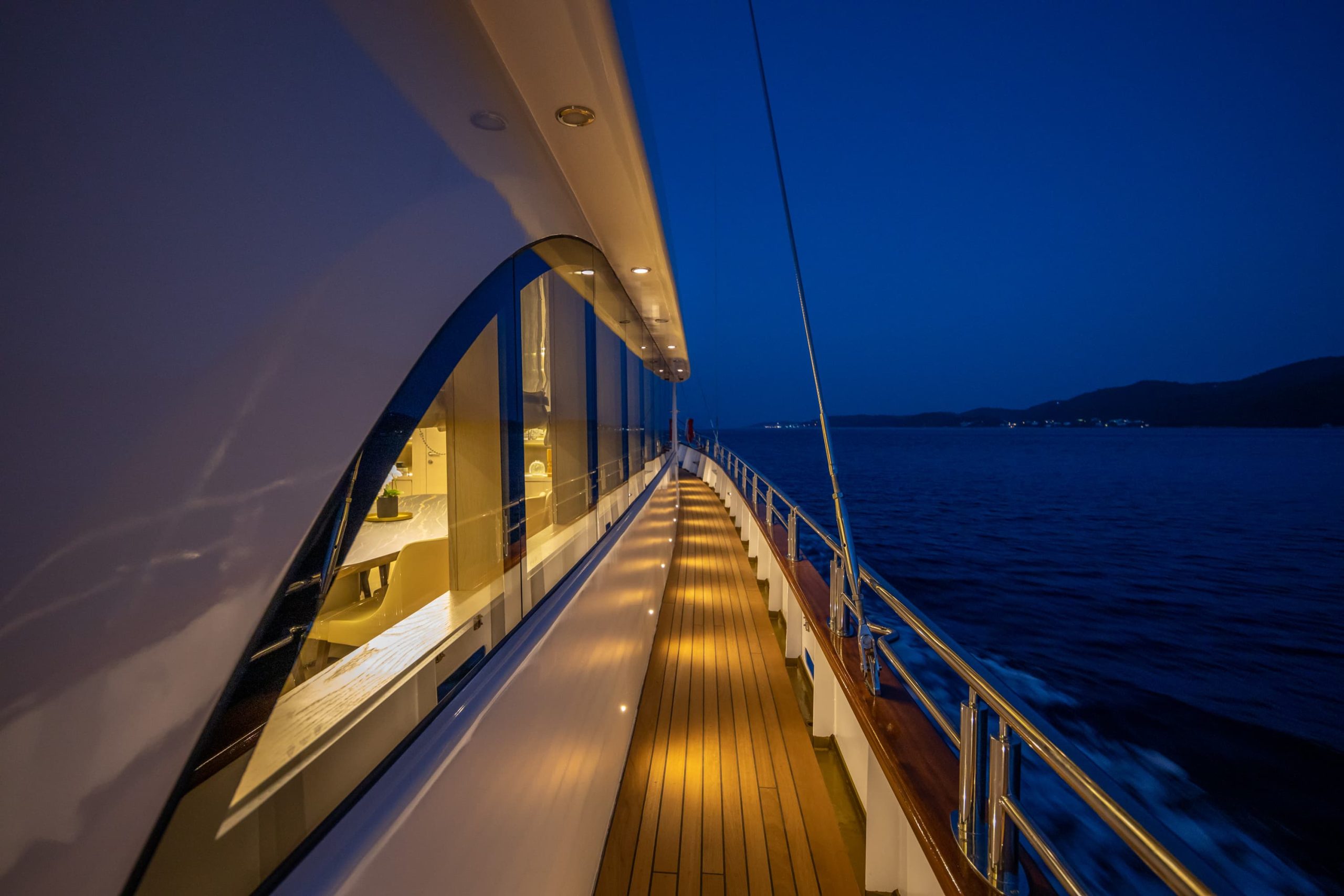 A yacht deck at night with warm lights illuminating the walkway, reflecting off the windows. The dark blue sea and distant shoreline are visible under a deep blue sky.
