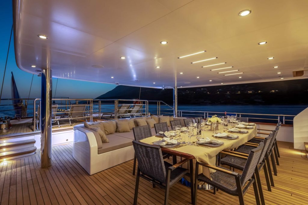 An elegant dining area on a yacht deck at dusk, featuring a long table set for dinner, cushioned seating, soft lighting, and a view of the ocean and distant coastline.